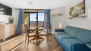 A modern living room with a blue sofa, wooden dining table, and chairs. There is a kitchen area with a wooden cabinet and a TV mounted on the wall. - Ocean View Ste: Hot Breakfast, 11th Floor (Myrtle Beach)
