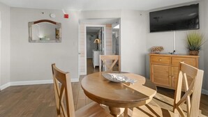 A dining area with a round wooden table, two wooden chairs, a wooden cabinet, and a flat-screen TV mounted on the wall. - Ocean View Ste: Hot Breakfast, 11th Floor (Myrtle Beach)