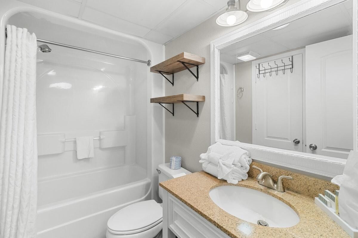 A modern bathroom with a bathtub, toilet, and sink. There are wooden shelves above the sink and a towel rack on the wall.
