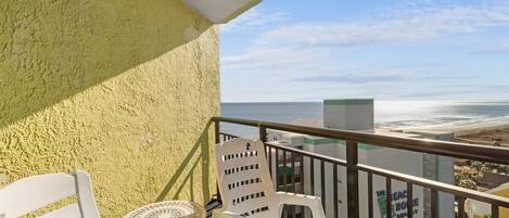 A balcony with white plastic chairs and a small round table, overlooking a beach and a building with a sign.
