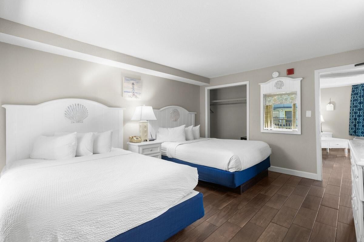 A hotel room with two beds, a wardrobe, a bedside table with a lamp, and a view of the outside.