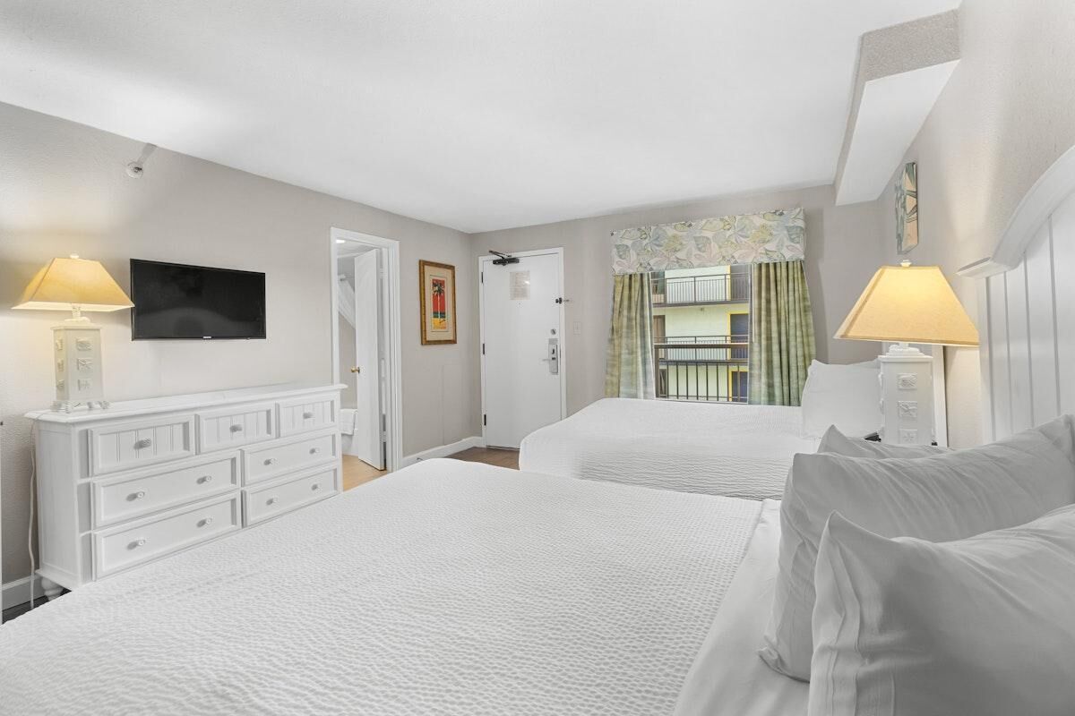 Une chambre d’hôtel avec deux lits, une commode, une télévision et un balcon avec des rideaux.