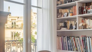 A room with a red chair, a bookshelf filled with books, and a view of buildings through large windows. - Marais Melody, by Veeve (Paris)