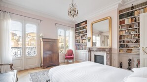 A bedroom with a large bed, a red chair, a bookshelf, and a fireplace. - Marais Melody, by Veeve (Paris)