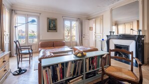 A spacious room with a large window, a fireplace, a sofa, a bookshelf, and a dining table. - Marais Melody, by Veeve (Paris)