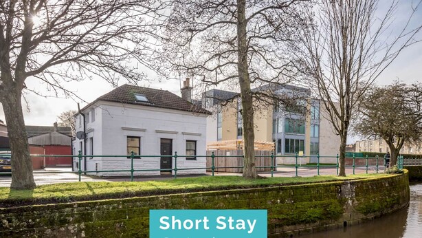 A white building with a black roof, a green fence, and a river in the foreground. - Kinnessburn Road Apartment (No 20) (St Andrews)