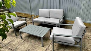 En uteplats med ett grått metallbord, en grå metallbänk och en grå metallsoffa med vita kuddar.