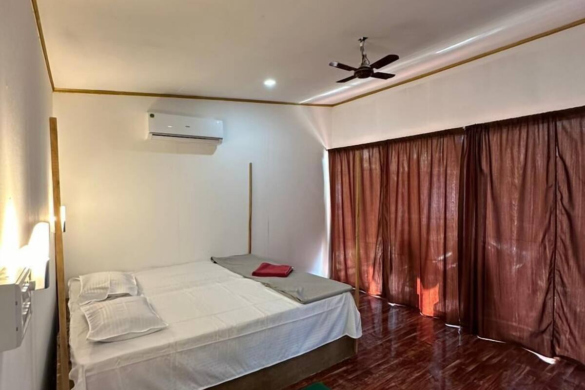 Um quarto com cama, ventilador de teto, ar condicionado e janela com cortinas.
