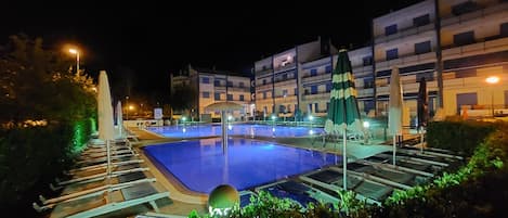 A swimming pool with lounge chairs and umbrellas, surrounded by a building with multiple balconies.