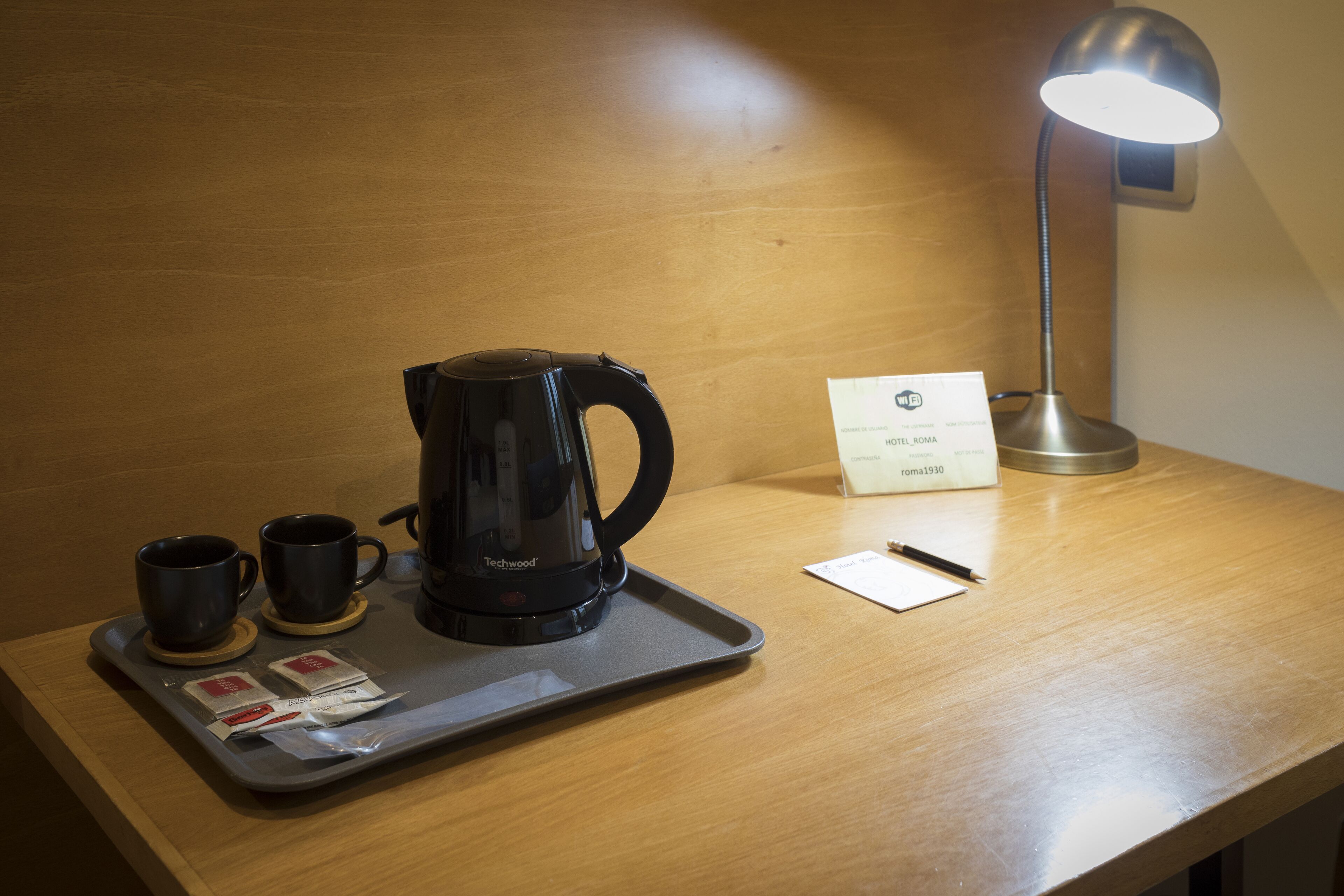 A hotel room with a wooden desk, a black coffee maker, two black cups, a tray with tea packets, a pen, a notepad, and a hotel sign.