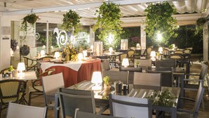 Un área de comedor al aire libre con mesas y sillas, plantas colgantes e iluminación ambiental.
