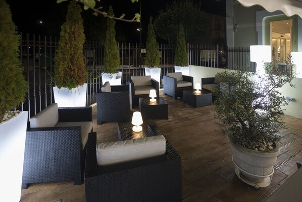 Un espacio al aire libre con mobiliario de mimbre y plantas en macetas, iluminado con luz tenue.