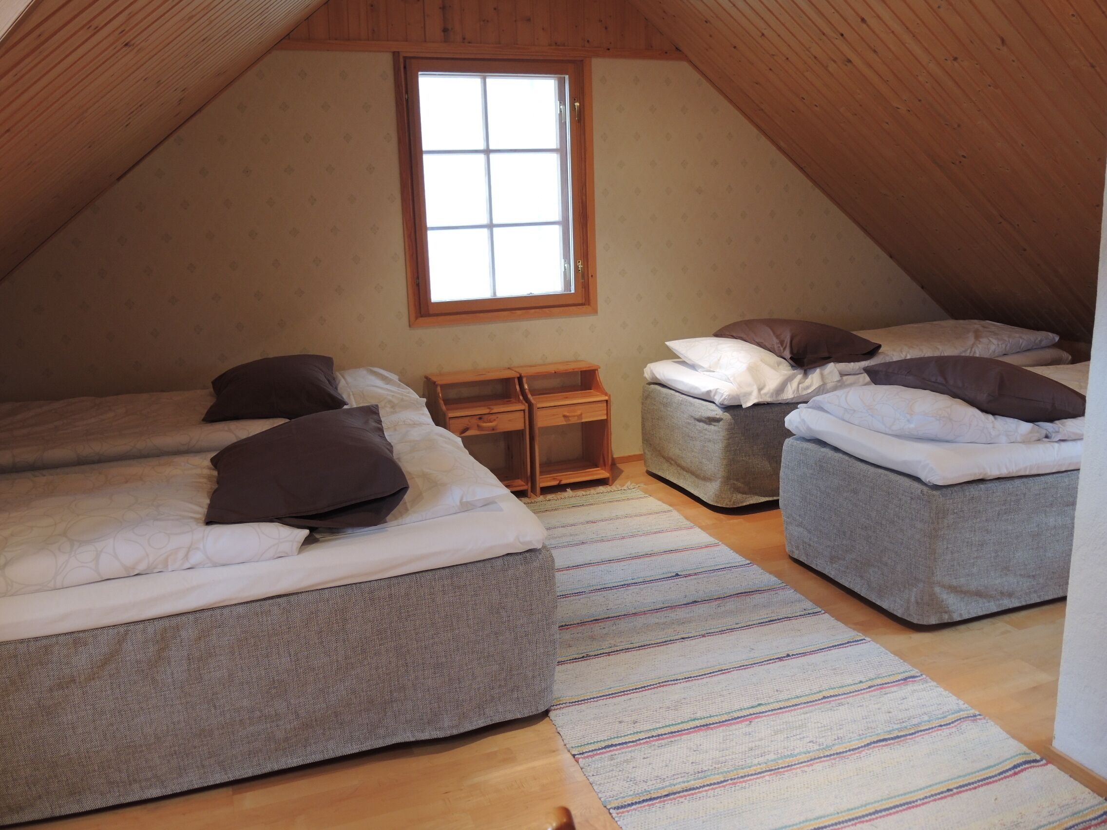 Un dormitorio abrigado en el ático, con dos camas, una mesita de noche de madera y una ventana que deja entrar luz natural.