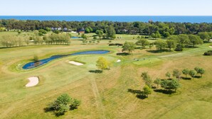 Luftaufnahme eines Golfplatzes mit Sandbunkern, einem Teich und einem bewaldeten Hintergrund.