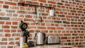 A kitchen with a brick wall, a coffee maker, a kettle, a toaster, and a white cup hanging on a rack. - Duplex 4pers - 100m du Métro - Smarttv - Fibre (Lille)