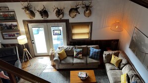 Un salon chaleureux avec un canapé, une table basse et une cheminée. On peut y admirer des têtes de cerf accrochées au mur, ainsi qu’une fenêtre équipée de stores.