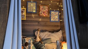 Une pièce chaleureuse et rustique, avec un plancher en bois, un lit recouvert d’une couette à rayures, un ours en peluche et une lanterne. Des guirlandes lumineuses et des tableaux encadrés décorent le mur.