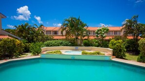 A swimming pool with a clear blue water, surrounded by lush greenery and a building with a red-tiled roof in the background. - Stylish, light villa w/pool. All modern amenities, gated. Right by the beach. (Rodney Bay)