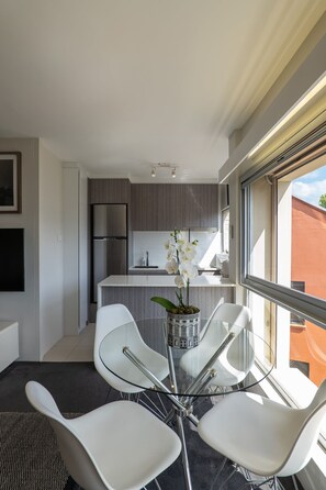 A modern kitchen with a stainless steel refrigerator, white cabinets, and a large window. There is a dining area with white chairs and a glass table.