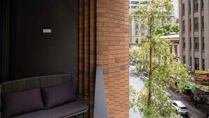 A balcony with a brick wall, a cushioned bench, and a view of a city street with buildings and a tree. - City Living at it's Finest (Sydney)