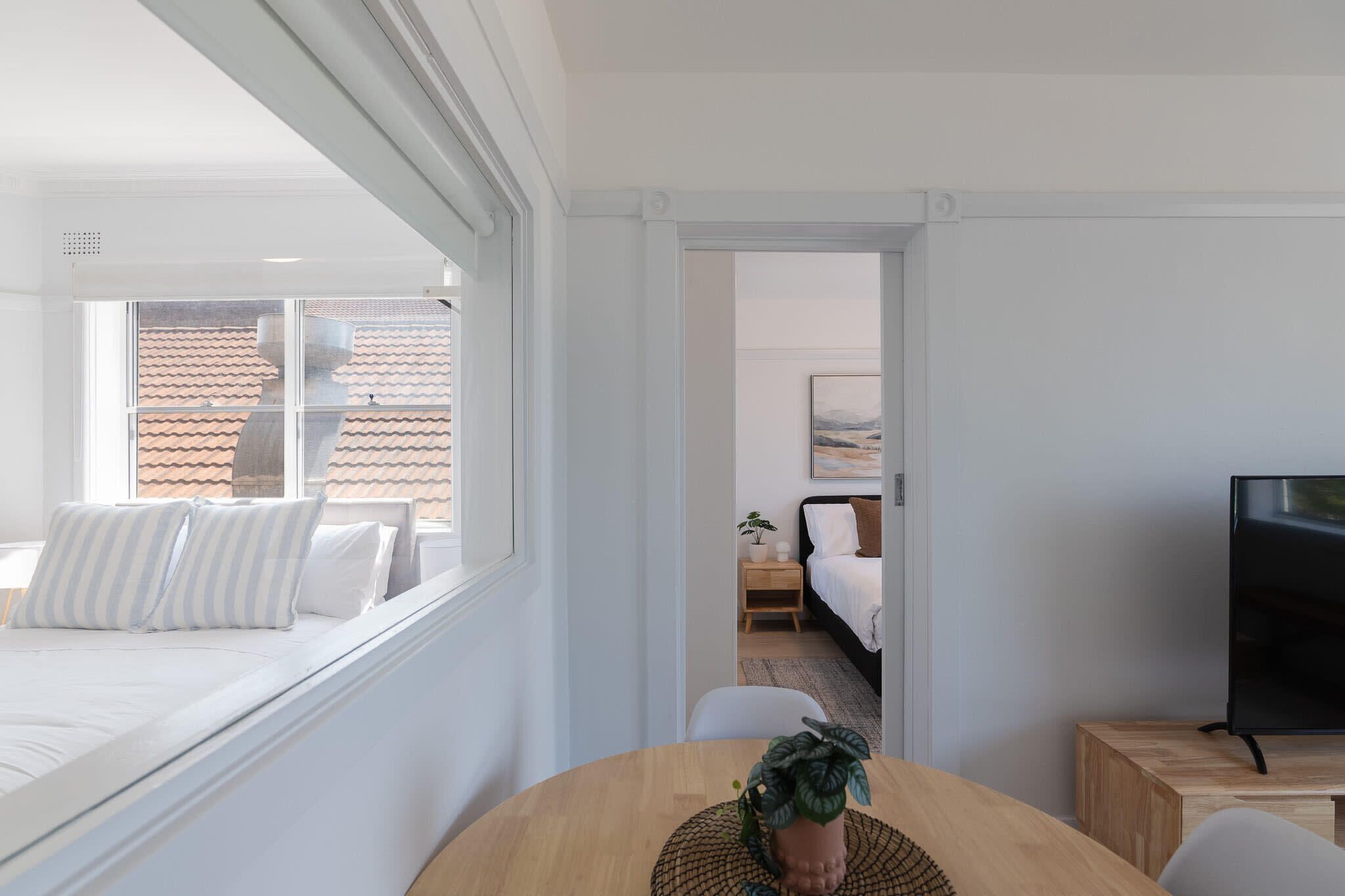 A modern hotel room with a bed, a TV, a dining table with a plant, and a view of the cityscape through large windows.