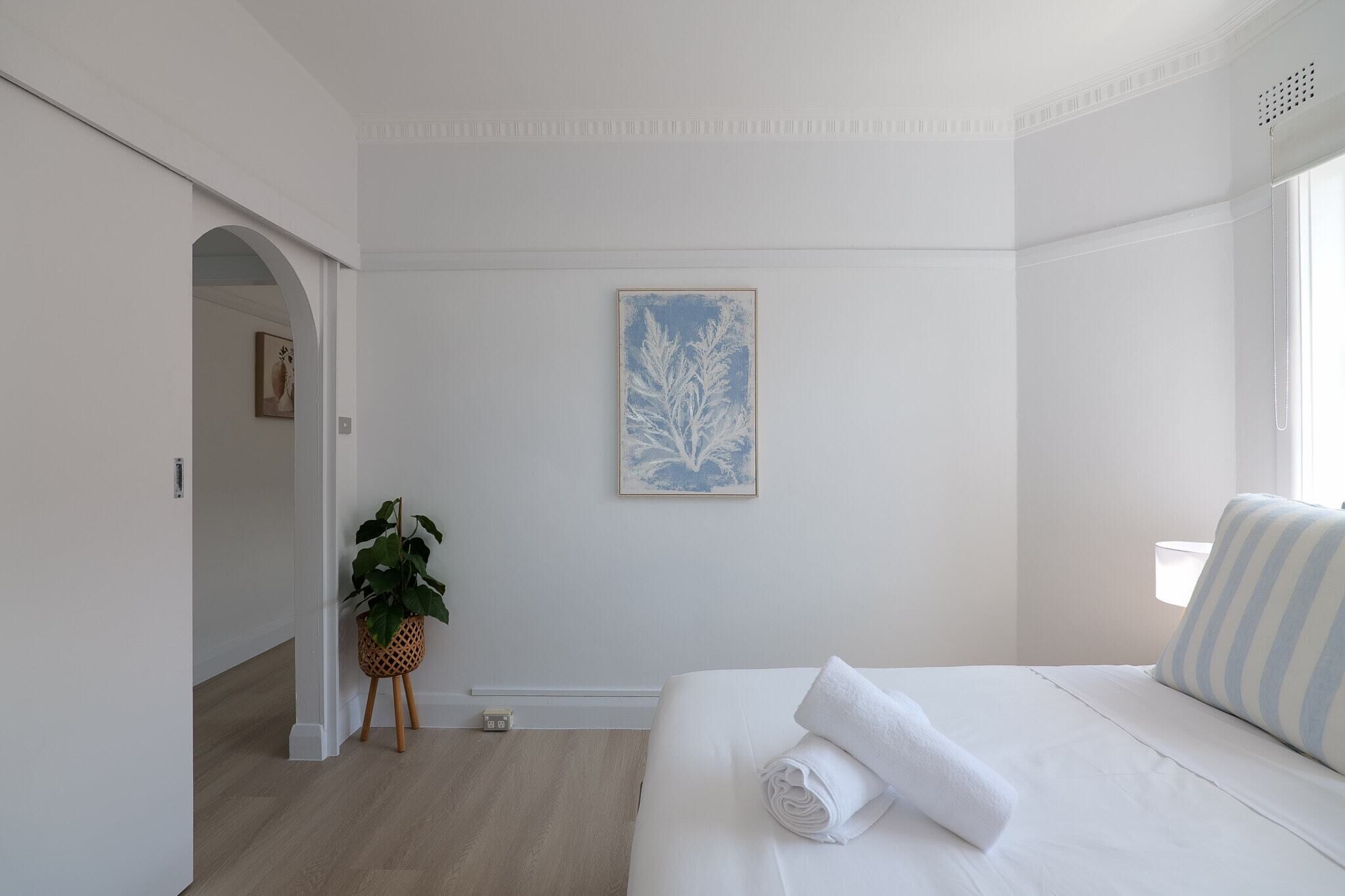 A minimalist hotel room with a bed, a framed artwork, a potted plant, and a view of a cityscape.