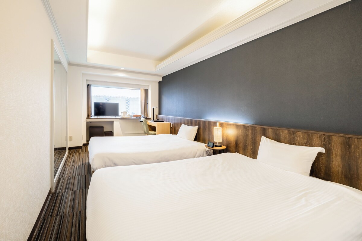 A hotel room with two beds, a desk, a TV, and a window.