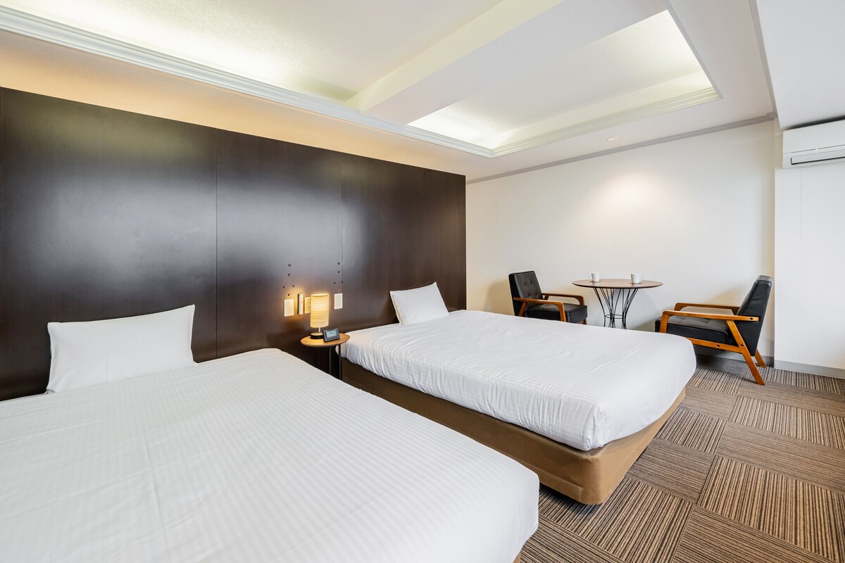 A hotel room with two beds, a desk, chairs, and a small table.
