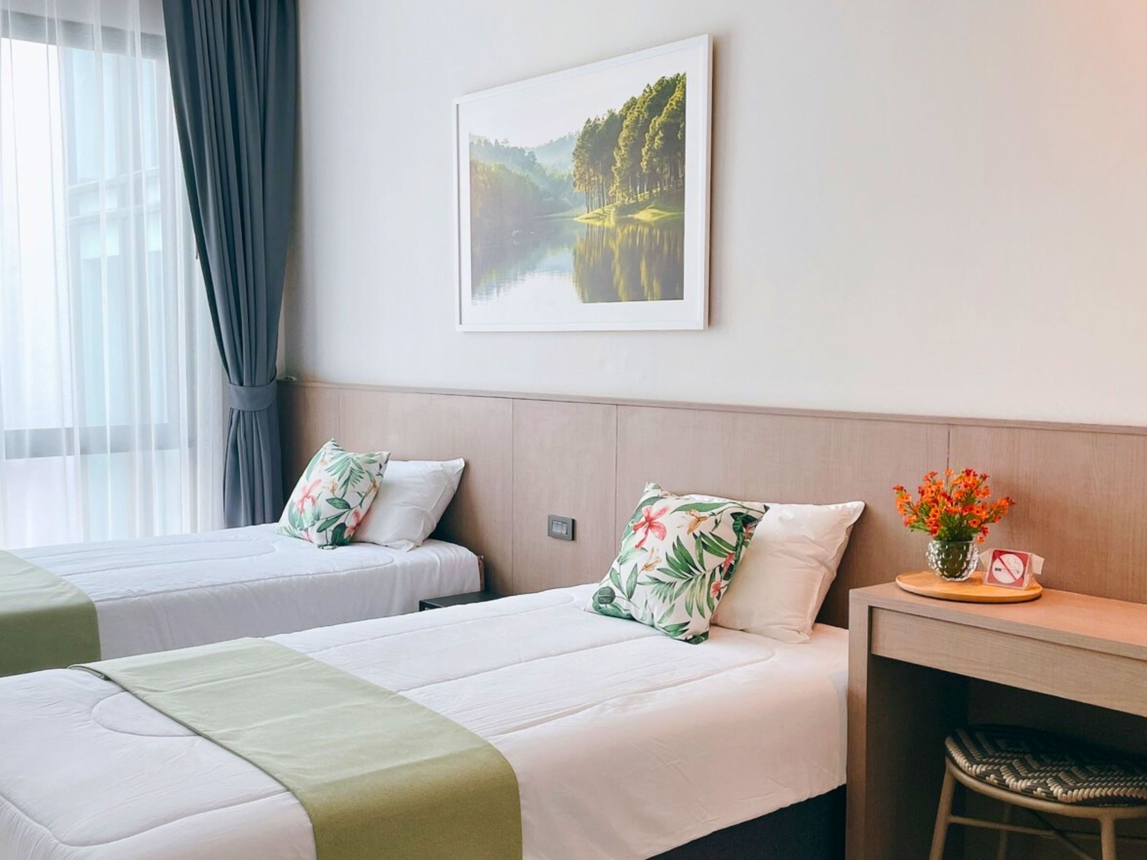 Habitación de hotel con dos camas, un cuadro enmarco de un paisaje, una mesita con un florero y una ventana con cortinas.