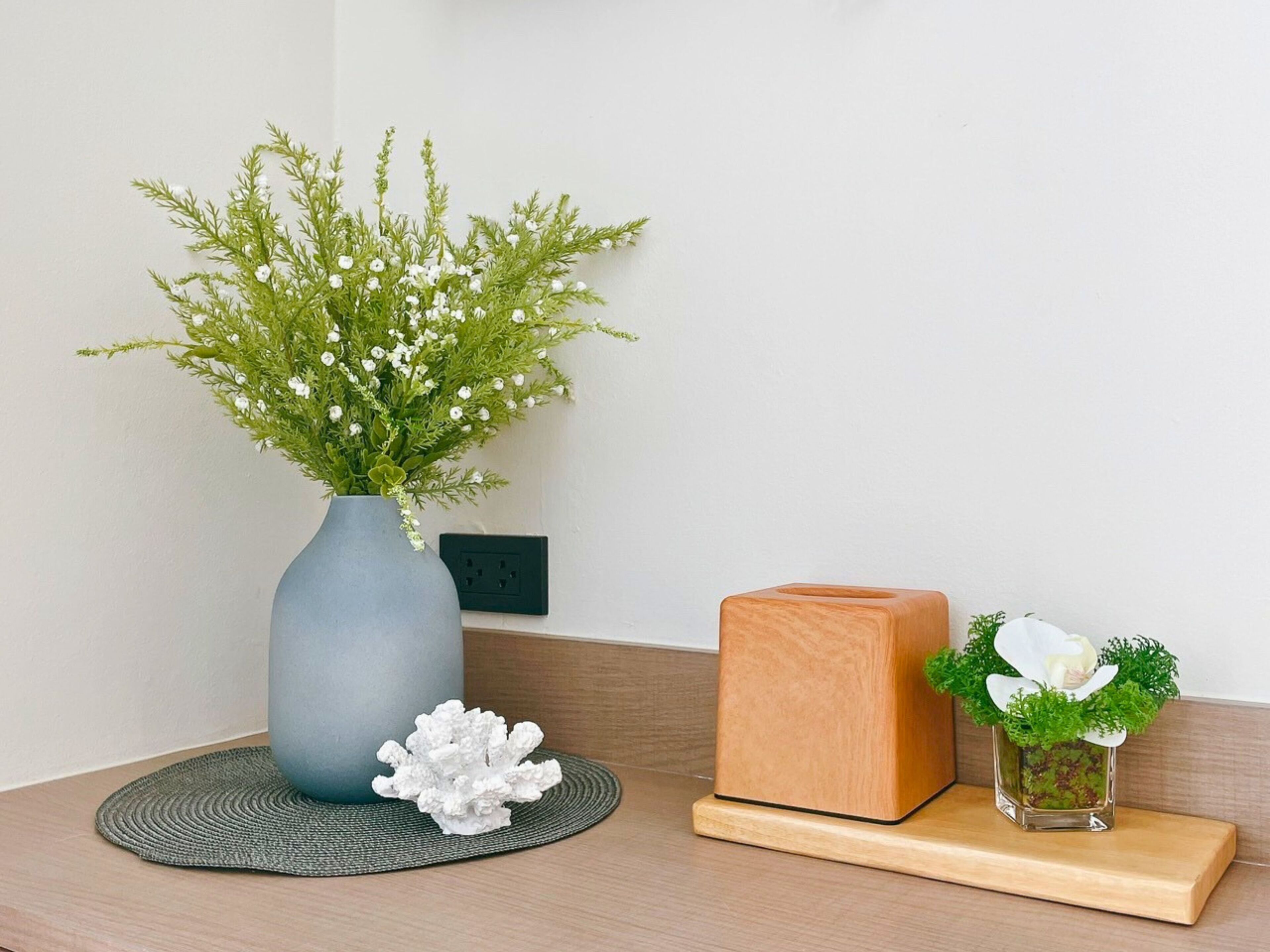 Une pièce dans laquelle on aperçoit un vase gris contenant des fleurs blanches et du feuillage vert, posé sur un tapis tissé, ainsi qu’un bloc de bois et un petit vase en verre avec des fleurs blanches, lui aussi placé sur une étagère en bois.