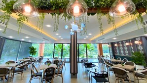 A modern dining area with multiple tables and chairs, a central column with hanging plants, and large windows offering a view of greenery outside. - Sand Dance Hotel (Prachuap Khiri Khan)