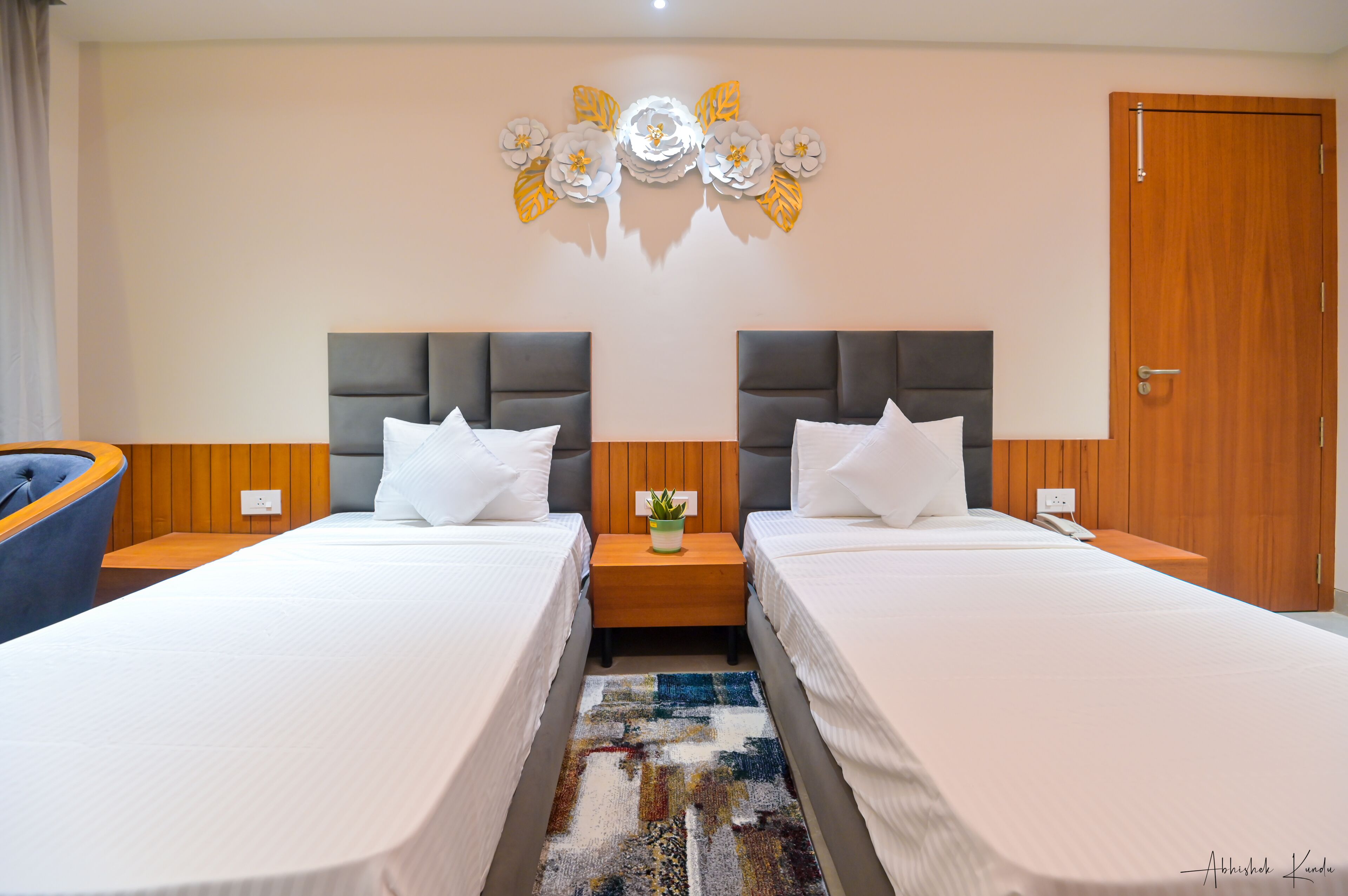 A hotel room with two beds, a wooden headboard, a nightstand with a plant, and a decorative wall piece.