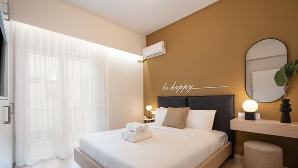 Quarto de hotel moderno com uma cama grande, uma mesa de cabeceira com abajur, um ar condicionado fixado na parede e uma frase motivacional na parede.
