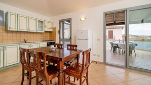 Een keuken met een eettafel en stoelen, een koelkast en uitzicht op een balkon met buitenmeubilair.