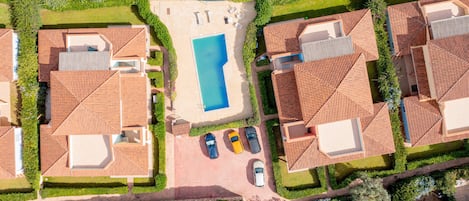 An aerial view of a residential area with a swimming pool, multiple houses with red-tiled roofs, and a few cars parked in the driveway.