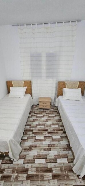 Una habitación con dos camas, piso de madera y una ventana con cortinas blancas.
