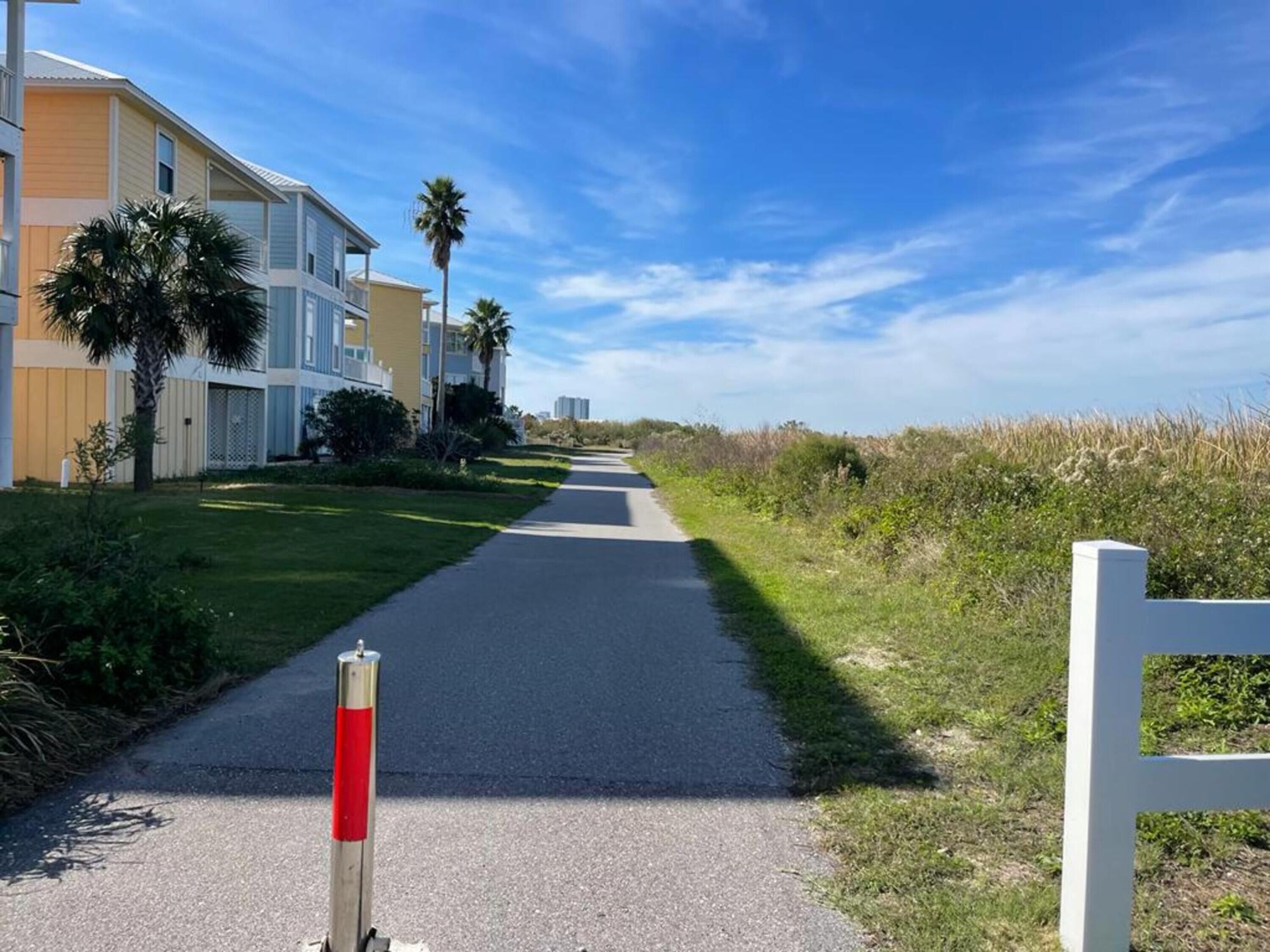A pathway leading to a residential area with modern buildings, a clear sky, and a grassy area with palm trees.