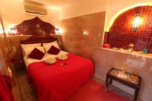 Una camera d'albergo con un letto, un tavolino con un vaso e una ciotola di frutta, e una parete con un arco decorativo e uno specchio.