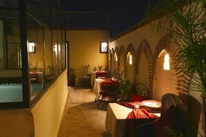 Area esterna con posti a tavola e sedie, illuminata da luci a parete, circondata da un muro di pietra con archi.