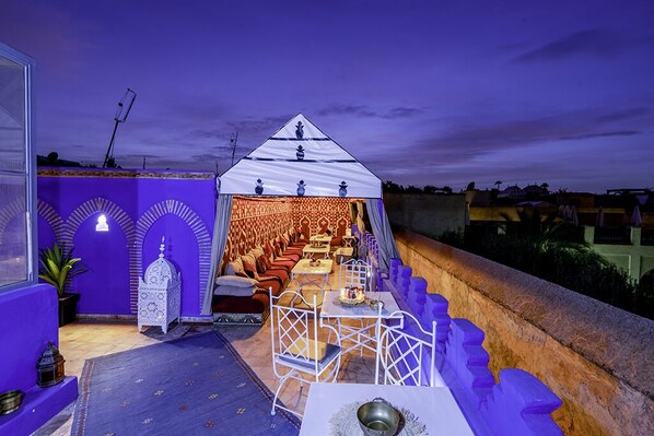 An outdoor seating area with a tent, tables, and chairs, surrounded by a purple wall and arches.