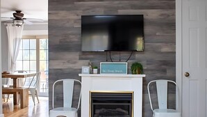 A modern living room with a flat-screen TV mounted on a wooden wall, a fireplace with a sign reading "Beach Sleep Beach," and white chairs. - Beach Daze: Cozy Sawyer Duplex, Fire Pit, 1 Mile to Warren Dunes (Sawyer)