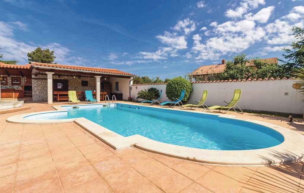 A swimming pool with a curved edge, surrounded by a tiled patio. There are several lounge chairs and a small building with a tiled roof.