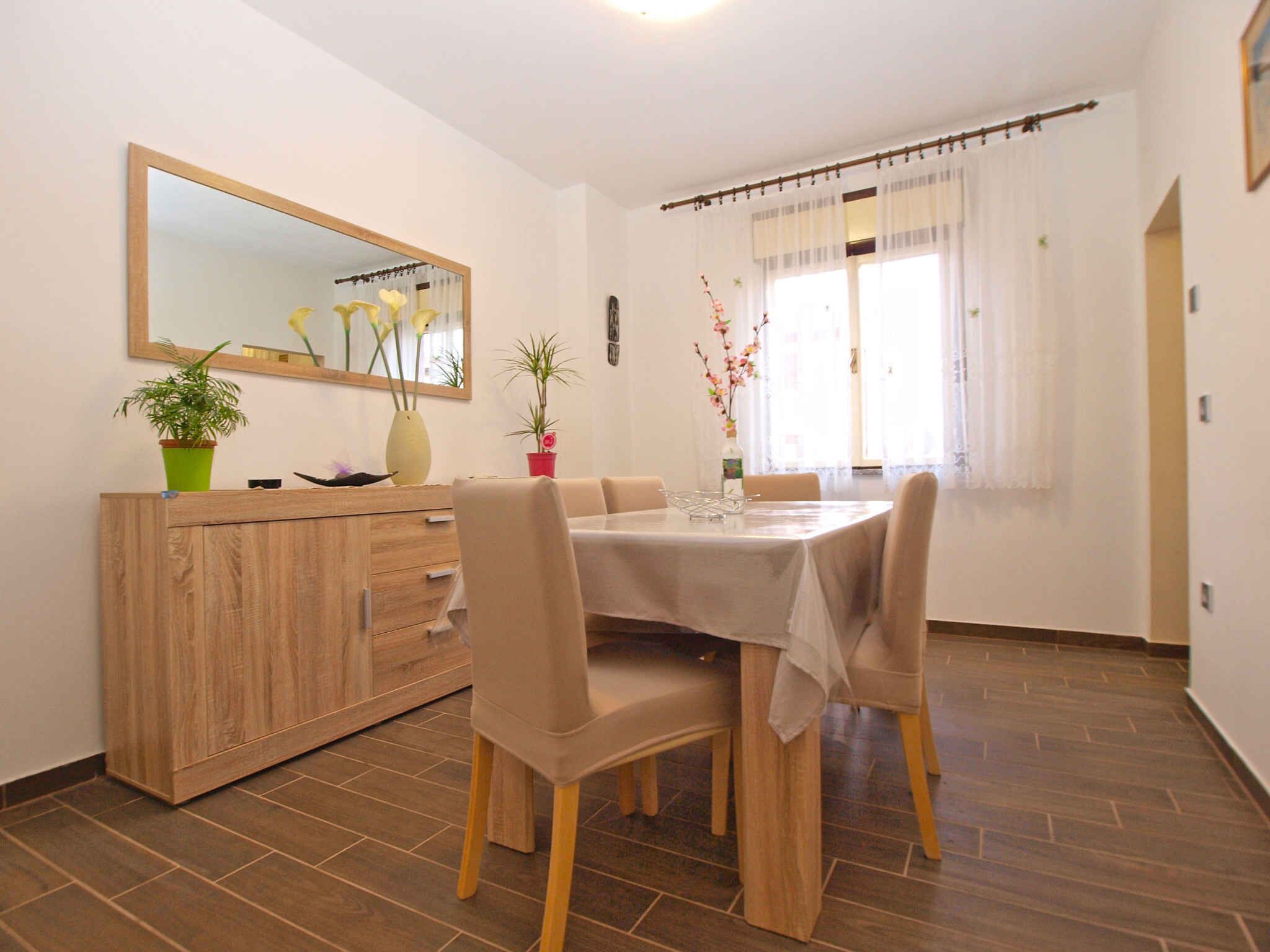 A dining area with a wooden cabinet, a round table, and four chairs. There is a mirror on the wall, and potted plants are placed on the cabinet.