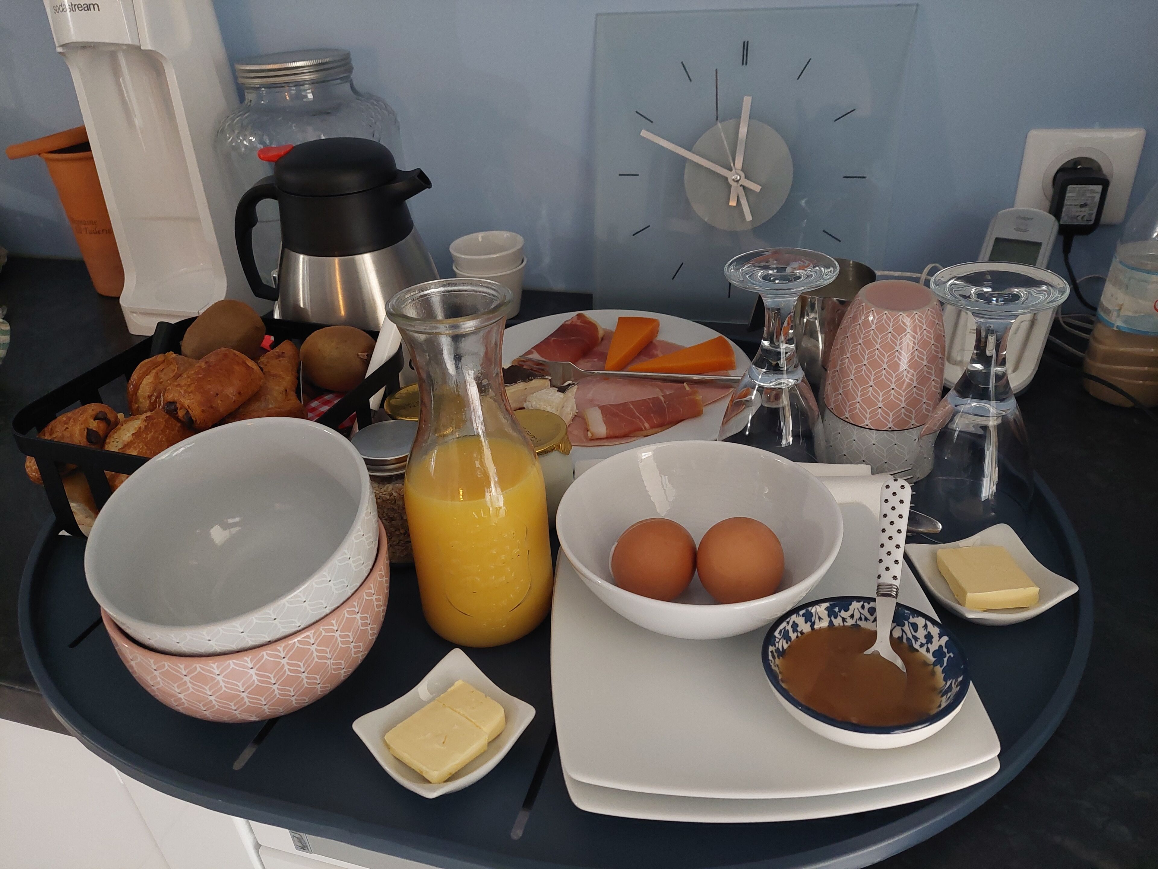 A breakfast table with various food items, a coffee maker, and a clock on the wall.