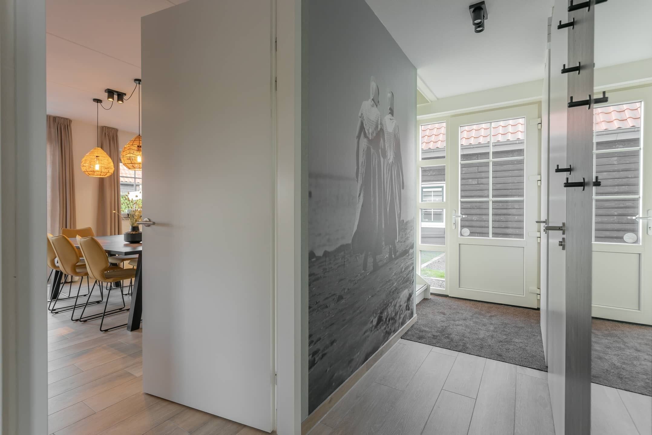A modern interior with a dining area featuring a table and chairs, a hallway with a large wall mural, and a door leading to an outdoor area with windows and a tiled roof.