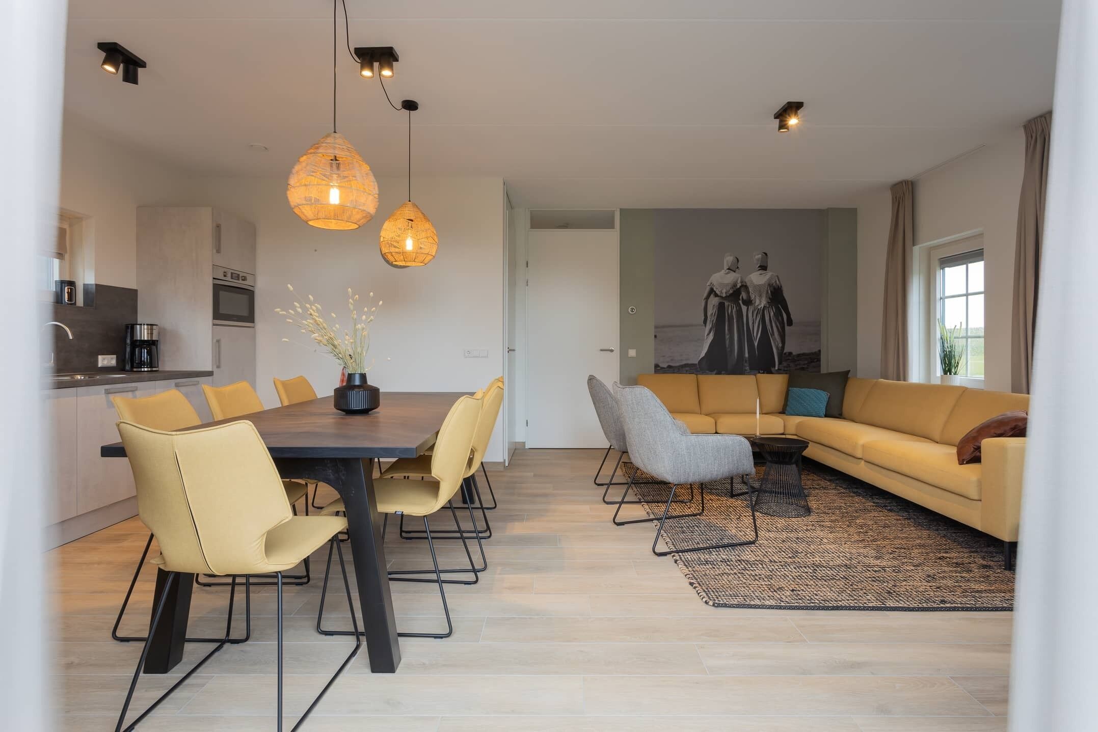 A modern living room with a sofa, dining table, and chairs. There is a kitchen area with appliances and a coffee machine.