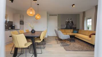 A modern living room with a sofa, dining table, and chairs. There is a kitchen area with appliances and a coffee machine.