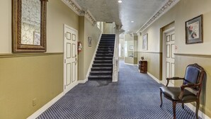 A hallway with a patterned carpet, a chair, a staircase, and a mirror with a decorative frame. - Cozy 2 bedroom Condo at the Château Sherbrooke, Blend of history & modern luxury (Montréal)