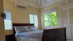 Un dormitorio con cama, dos ventanas con contraventanas, un equipo de aire acondicionado y una mesita de noche.