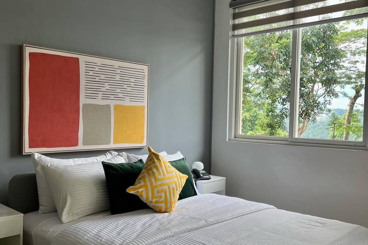 A neatly arranged bedroom with a bed, pillows, a bedside table, and a window with a view of trees.